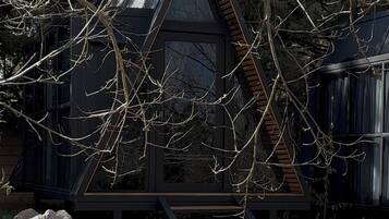 Una moderna baita a forma triangolare con una scala in legno che conduce a un ingresso con porta in vetro, immersa tra alberi e un'area verde.