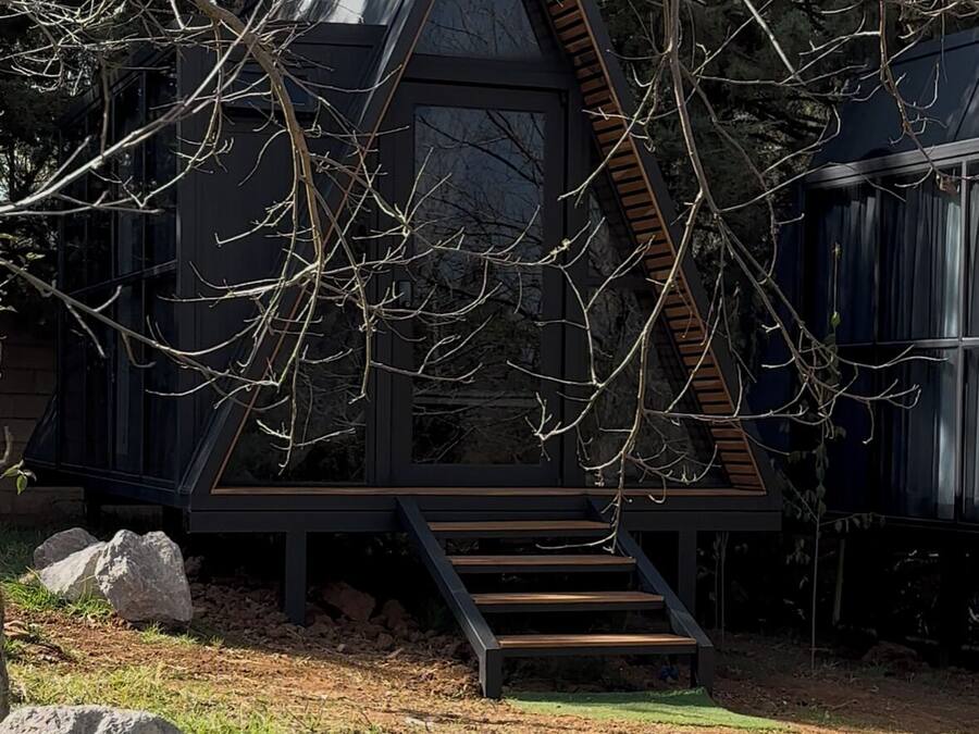 Una moderna baita a forma triangolare con una scala in legno che conduce a un ingresso con porta in vetro, immersa tra alberi e un'area verde.