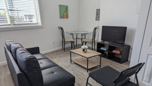 A modern living room with a black leather sofa, a small round table with a vase of flowers, a TV stand with a flat-screen TV, and a window with blinds. - Charming 1-bedroom apartment in welcoming Pittsburgh with AC (Pittsburgh)