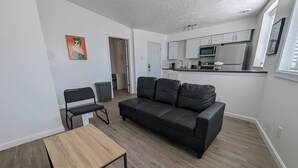 A modern living room with a black leather sofa, a wooden coffee table, and a black chair. A kitchen with white cabinets and a microwave is visible in the background. - Charming 1-bedroom apartment in welcoming Pittsburgh with AC (Pittsburgh)