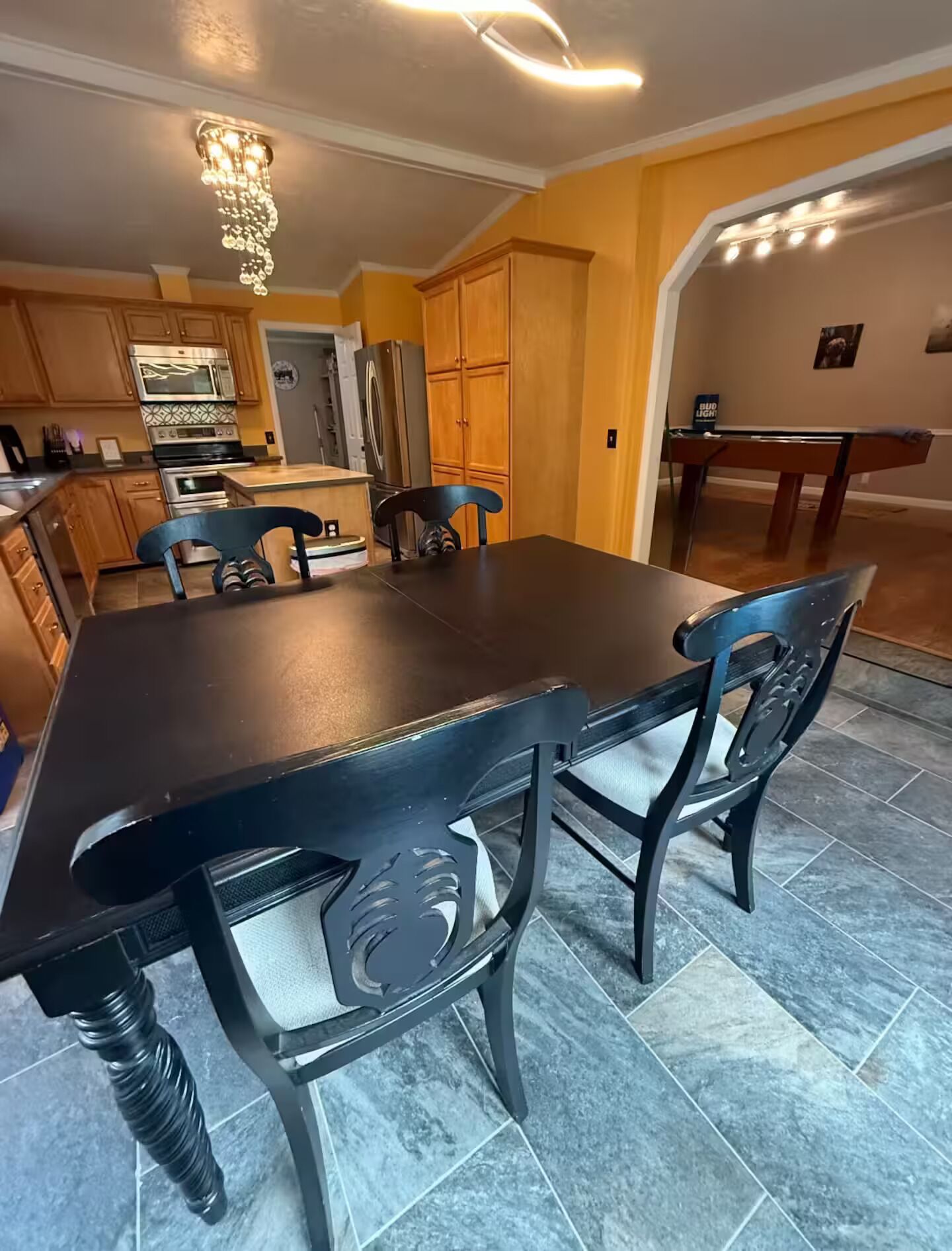A dining area with a dark wooden table and chairs, a chandelier, and a kitchen in the background.
