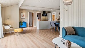 A modern living room with a blue sofa, a small round table, and a wooden floor. There is a kitchen area with a blue cabinet and a dining table with chairs. - 5 Star Holiday Home in Hals (Hals)