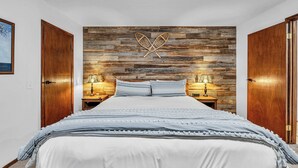 Un dormitorio con panel de pared de madera, una cama con ropa de cama a rayas, dos mesitas de noche con lámparas y un cuadro enmarcado en la pared.