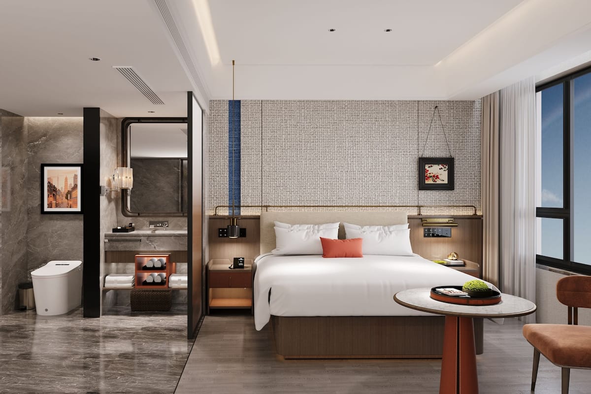 Ein modernes Hotelzimmer mit einem großen Bett, einem kleinen Tisch, einem Sessel, einem Badezimmer mit Dusche und Blick auf die Stadt durch das Fenster.