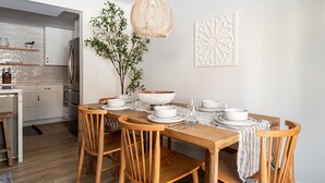 A dining area with a wooden table set for four, surrounded by wooden chairs. A woven pendant light hangs above, and a potted plant is on the table. The room has white walls and a modern kitchen in the background.