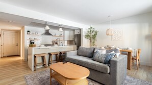 A modern living room with a grey sofa, wooden coffee table, and a kitchen area with white cabinets and stainless steel appliances.