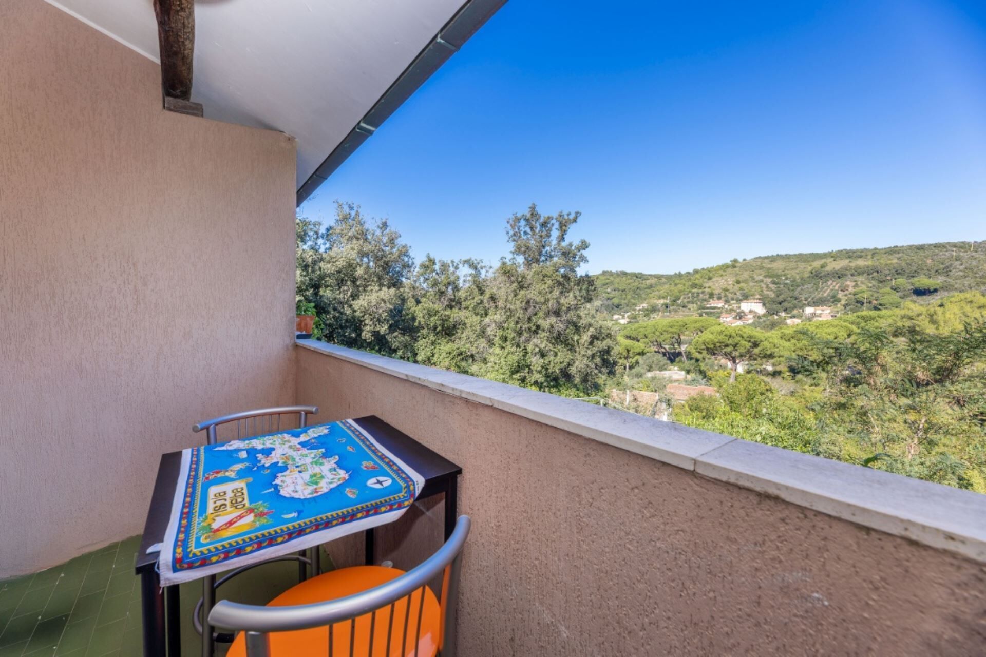 A balcony with a table and chair, overlooking a green landscape with trees and buildings.