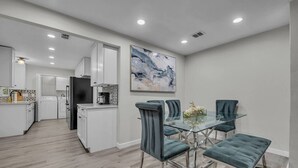 A modern kitchen with white cabinets, a black refrigerator, and a coffee maker. There is a dining area with a glass table and four blue chairs.