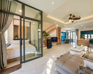A modern living room with a sofa, coffee table, and TV. There is a staircase leading to a bedroom with a bed. The room has a ceiling fan and recessed lighting. - The Moon Villa - Novaworld Phan Thiet (Tien Thanh)