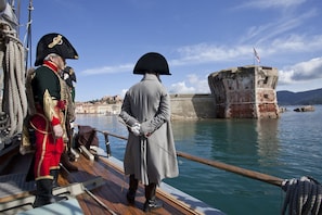 A historical ship with two individuals in period attire, a wooden deck, and a stone structure in the background.