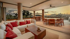 A spacious living area with a sofa, coffee table, and dining set. A large window offers a view of the ocean and palm trees.