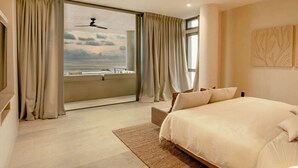 A bedroom with a large bed, a view of the sea through a balcony, and a ceiling fan.