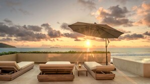 A sunlit outdoor seating area with lounge chairs and an umbrella, overlooking a beach and ocean at sunset.