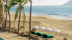 A beachside pool area with lounge chairs, umbrellas, and palm trees.