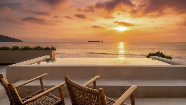 An infinity pool with a view of the sunset over the ocean, two wicker chairs, and a small island in the distance.
