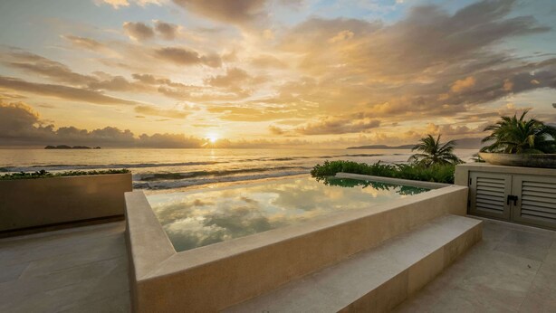 Une piscine offrant une vue imprenable sur le coucher de soleil sur l’océan, bordée de palmiers et dotée d’une architecture moderne.