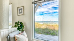 Ein Zimmer mit großem Fenster mit Blick auf einen Strand, ein Sessel mit Kissen und ein gerahmtes Bild an der Wand.