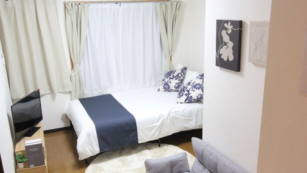 A bedroom with a bed, a grey sofa, a white rug, and a TV on a wooden stand. - Small-Town Feel, Big-City Access | Homey Stay in Nakano (中野区野方)