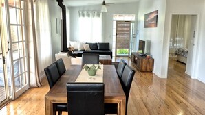 Uma sala de estar ampla com uma mesa de jantar de madeira, cadeiras de couro preto, um sofá preto e uma televisão sobre um suporte de madeira.
