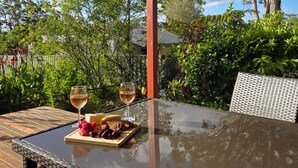 Uma mesa de vidro com duas taças de vinho, uma tábua de madeira com frutas e queijo, e uma cadeira de vime.