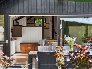 A modern outdoor seating area with wicker furniture and a wooden table, leading to a kitchen with white cabinets and a television. - 5 Star Holiday Home in Nykobing Sj (Nykobing Sjaelland)