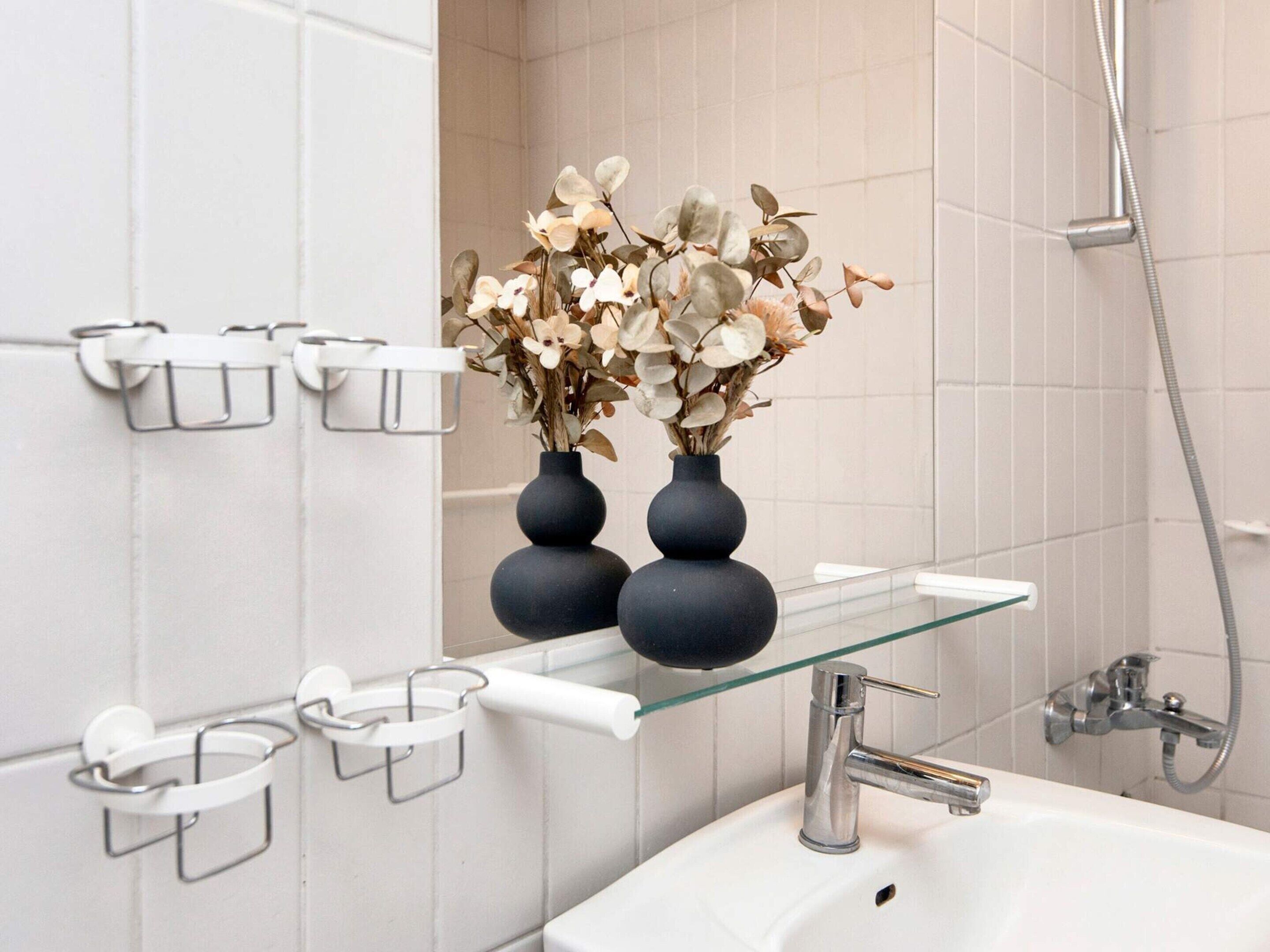 Un baño con un lavamanos blanco, dos jarrones negros con flores secas y un estante de vidrio con sujetatelas blancos.