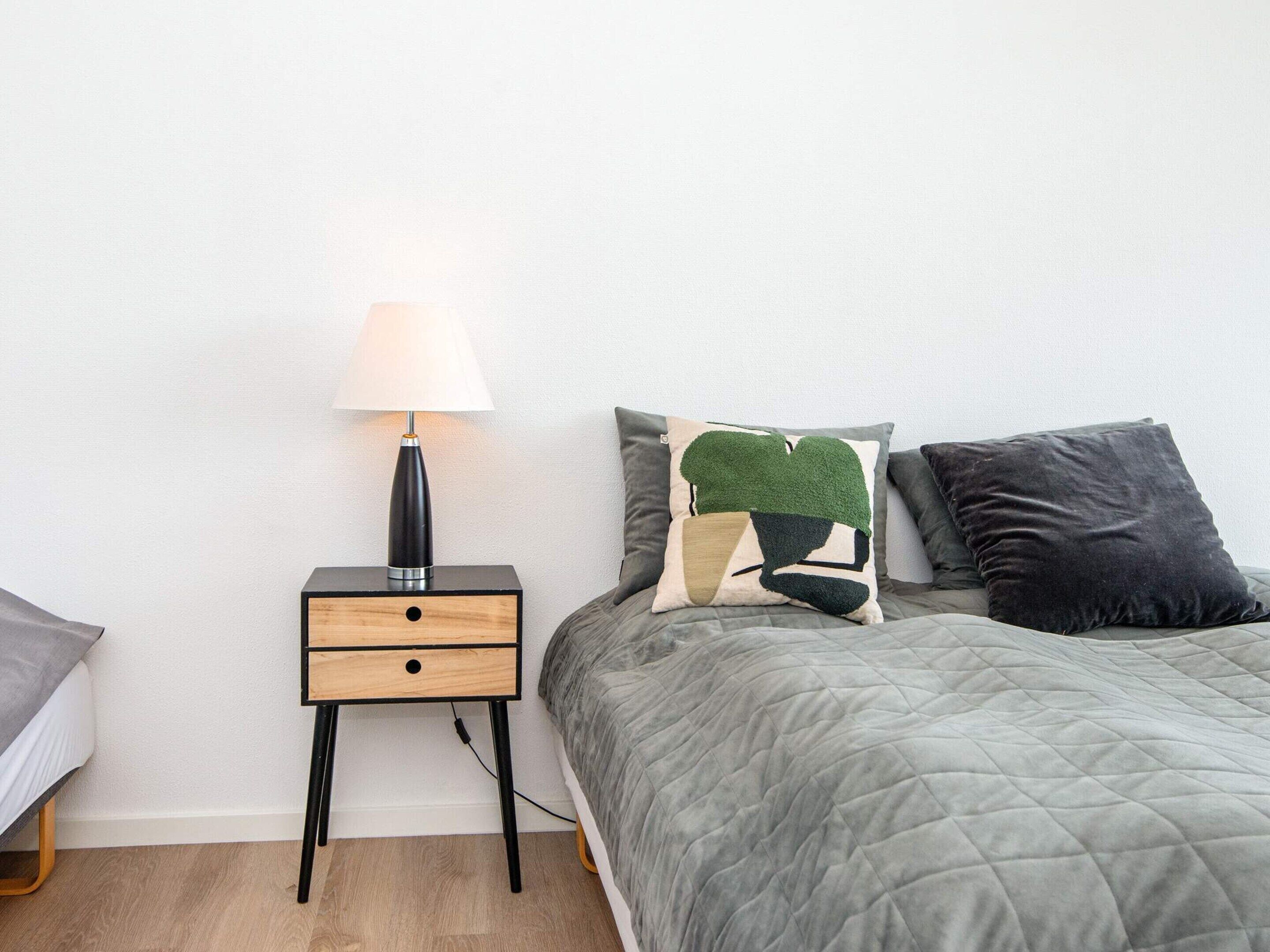 Ein ordentlich eingerichtetes Schlafzimmer mit einem Bett, einem Nachttisch mit Lampe und einem kleinen Holzbeistelltisch mit Schubladen.