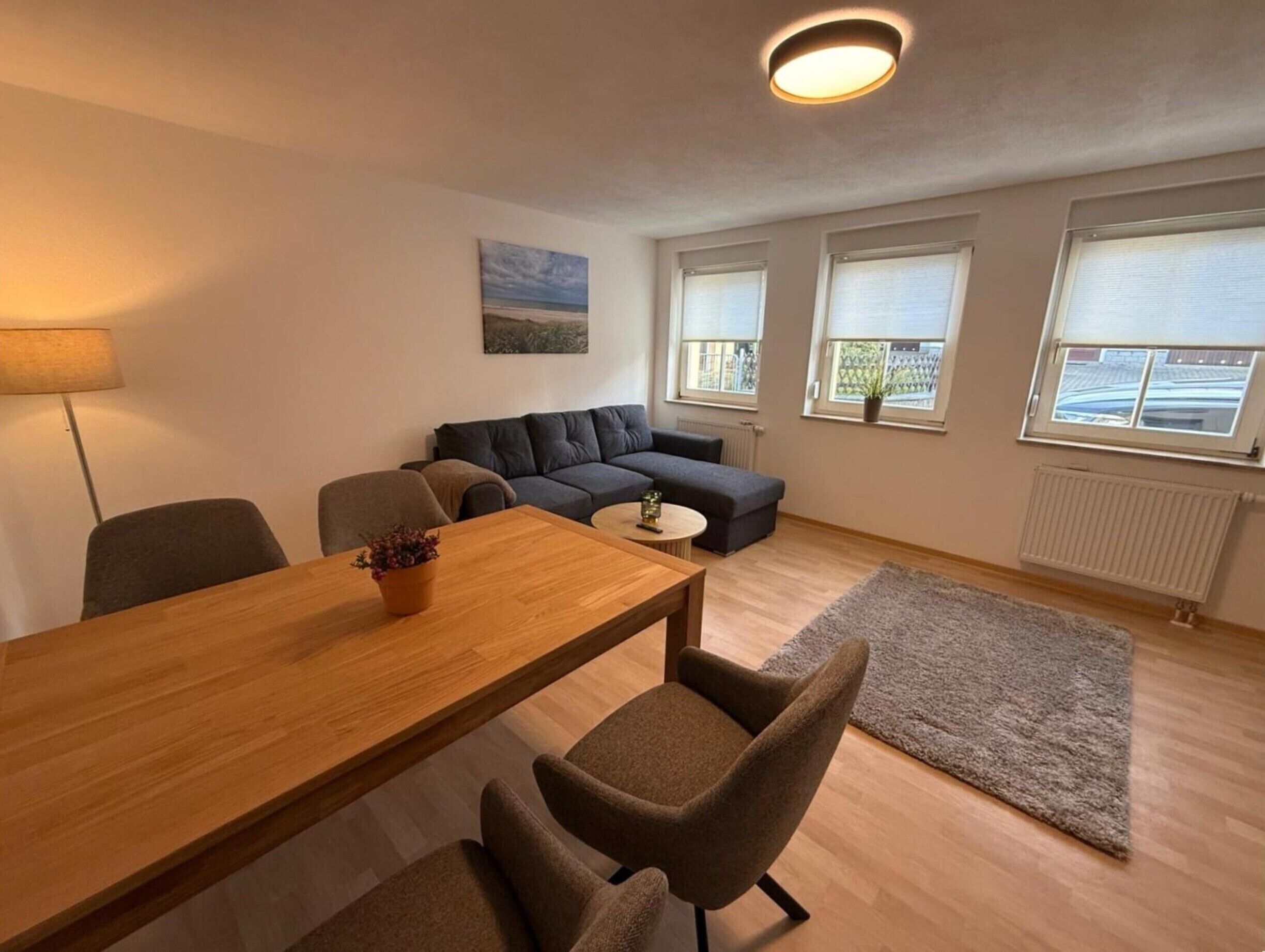 Uma sala de estar moderna com uma mesa de jantar de madeira, duas poltronas, um sofá cinzento e uma pequena mesa lateral. Há três janelas com persianas, um radiador e um quadro na parede.