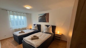 Ett hotellrum med två sängar, ett fönster med gardiner, en tavla på väggen och ett nattduksbord med en lampa.