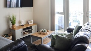 Een moderne woonkamer met een flatscreen televisie, een houten salontafel, een bank en uitzicht op de omgeving door grote ramen.