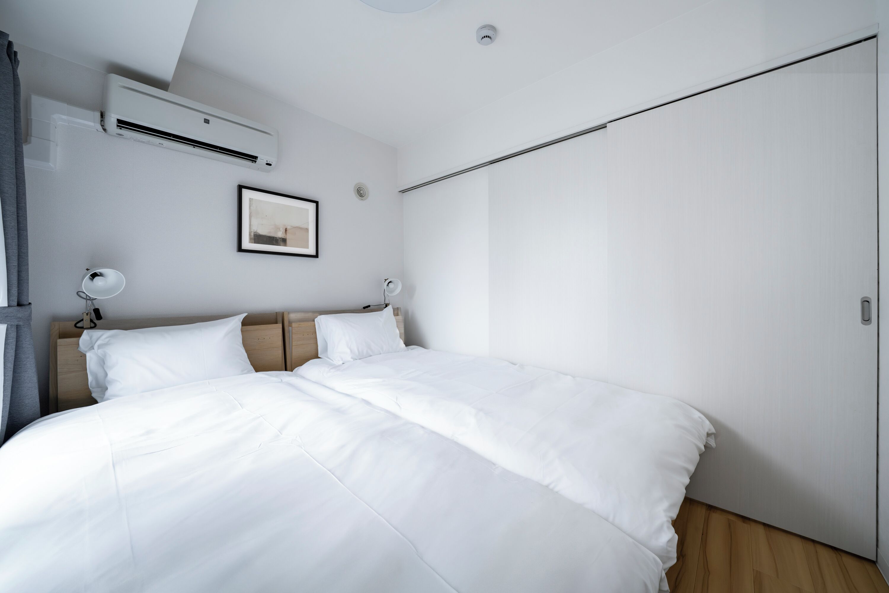 A modern bedroom with a large bed, white bedding, a ceiling-mounted air conditioner, a bedside table with a lamp, and a framed picture on the wall.