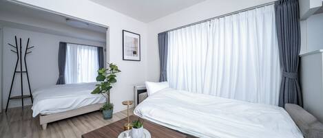 A modern hotel room with two single beds, a wooden coffee table with decorative items, a potted plant, and a large window with curtains.