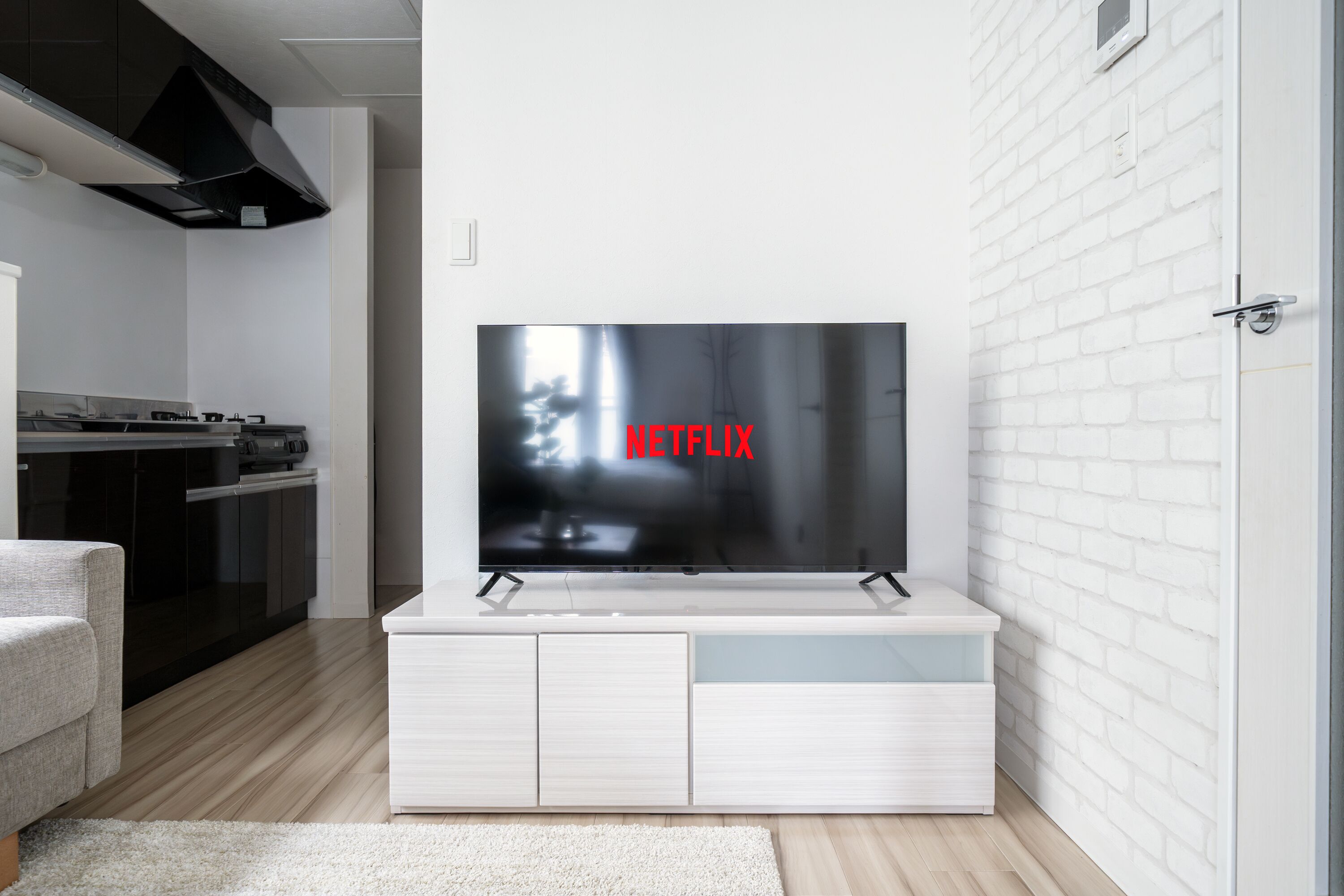Ein modernes Wohnzimmer mit einem Flachbildfernseher an einem weißen Schrank, einem Küchenbereich mit dunklen Schränken und einem Herd, und einer Wand aus weißen Ziegelsteinen.