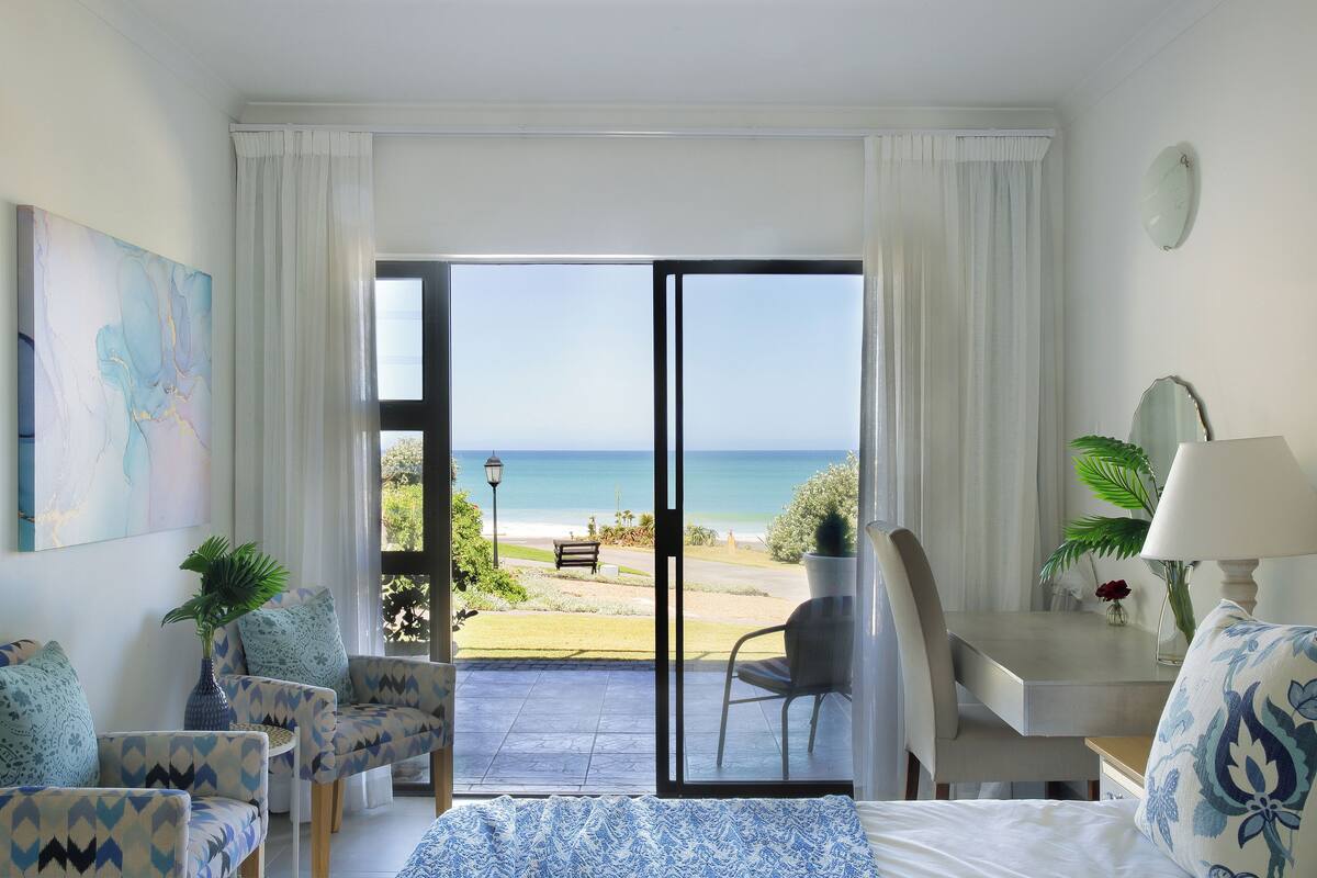 Habitación de hotel con cama, dos sillones, un escritorio con lámpara y vista a la playa.