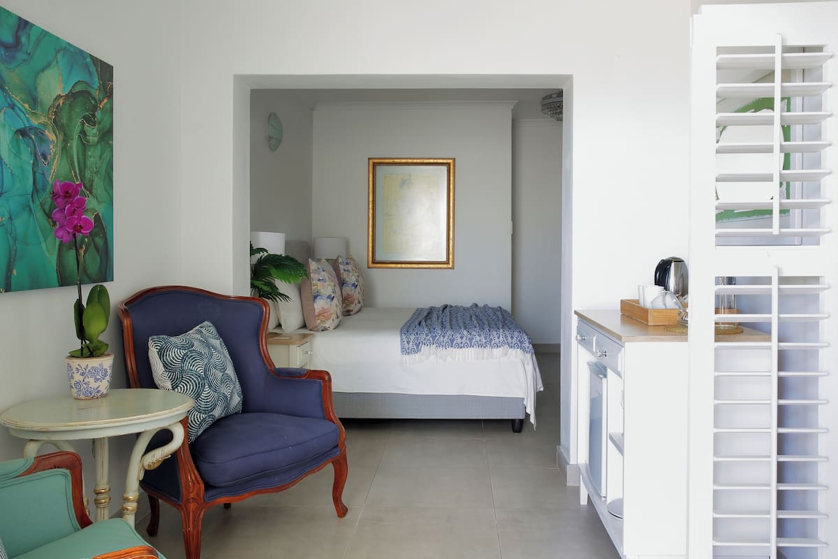Un dormitorio con cama, una silla, una mesita, una planta en maceta y un armario blanco con estantes.