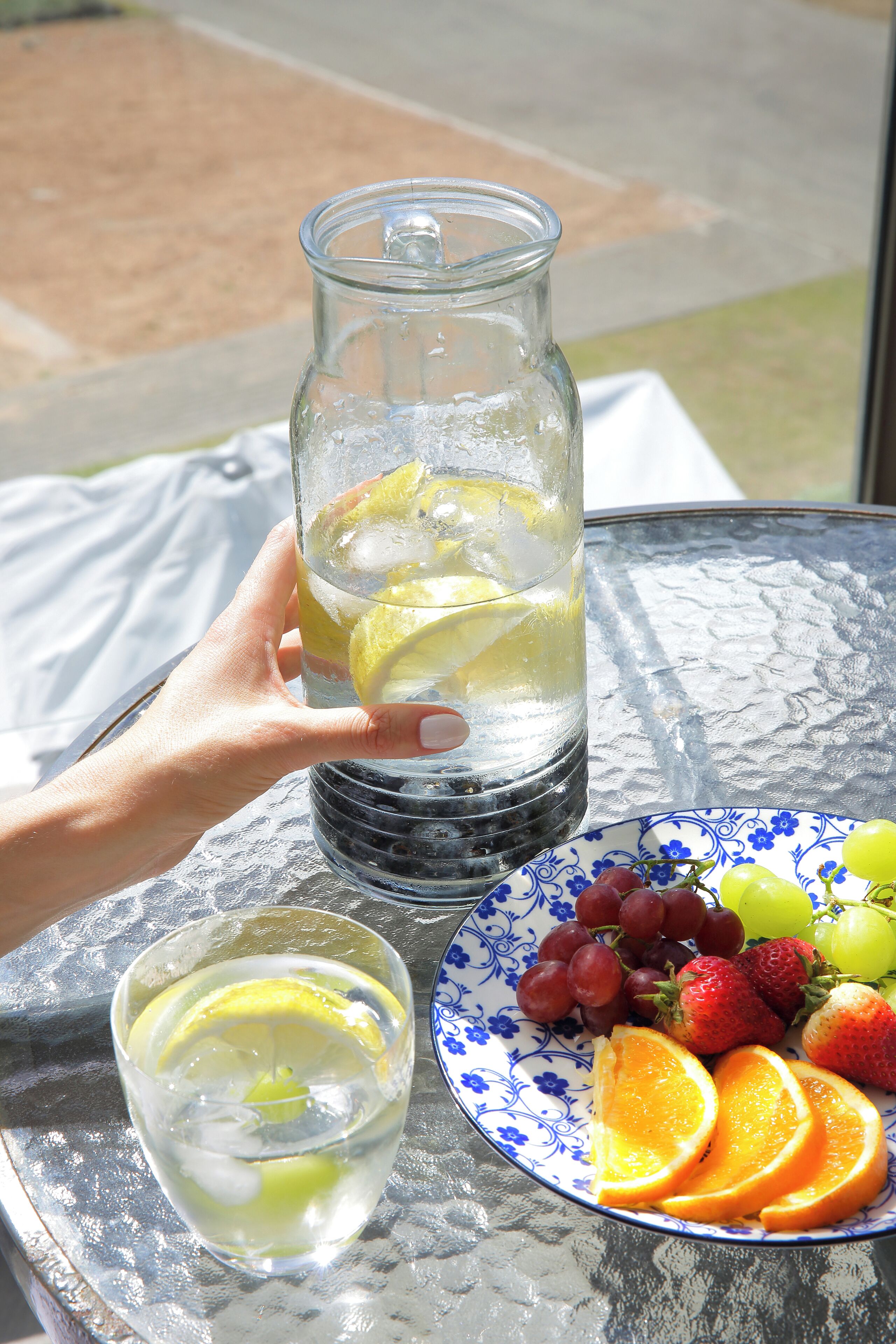 A hand holding a glass pitcher with ice and lemon slices, a glass of water, and a plate with grapes, strawberries, and orange slices on a table.