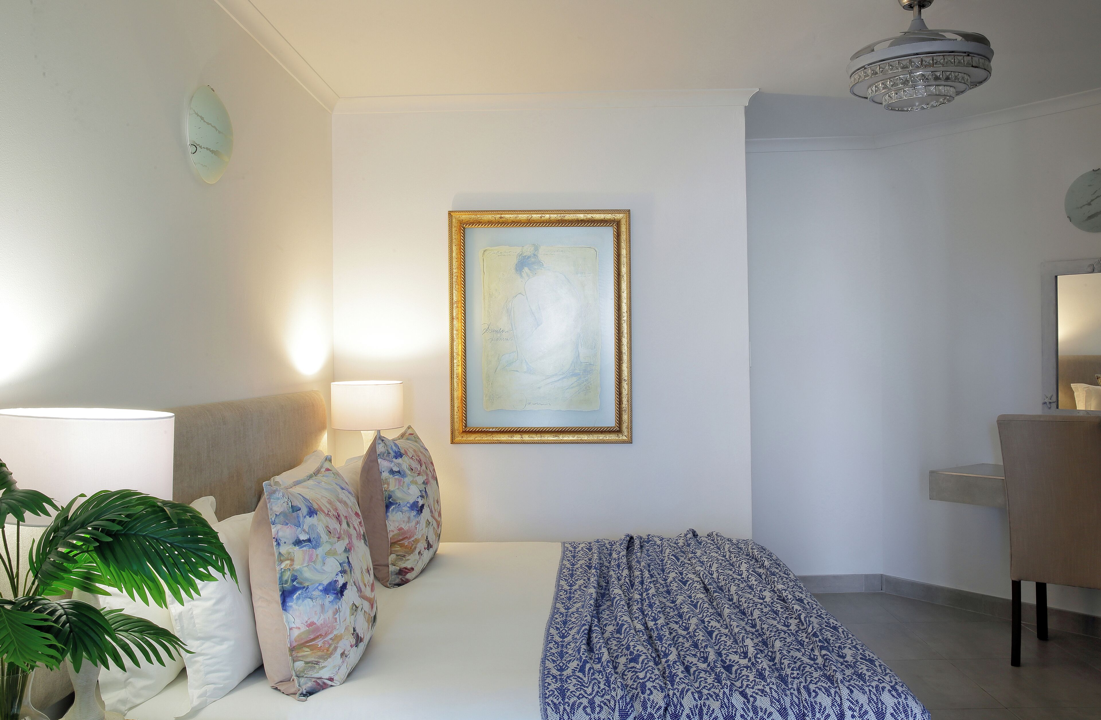 A hotel room with a bed, a desk, a chair, a lamp, a plant, and a framed artwork on the wall.
