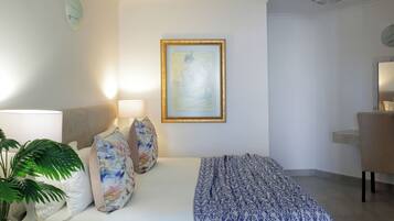 A hotel room with a bed, a desk, a chair, a lamp, a plant, and a framed artwork on the wall.
