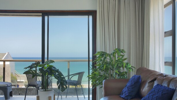 A living room with a large sliding glass door leading to a balcony with a view of the sea. There is a brown leather sofa with blue cushions, a white coffee table, and two chairs. A large potted plant is in the corner.