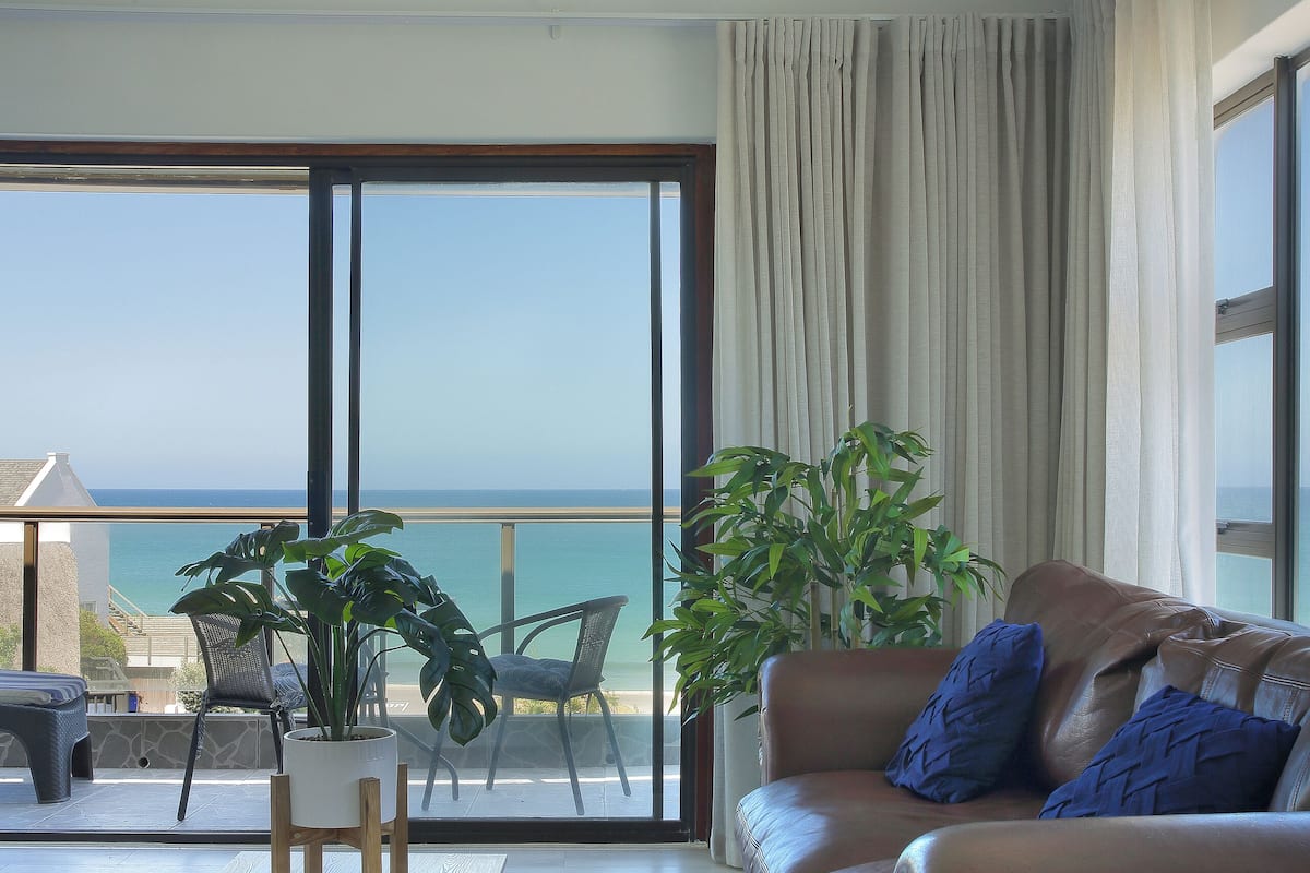 Un salón con un gran ventanal corredera que da a un balcón con vistas al mar. Hay un sof á de cuero marrón con cojines azules, una mesa de centro blanca y dos sillones. Una planta grande en maceta está en la esquina.