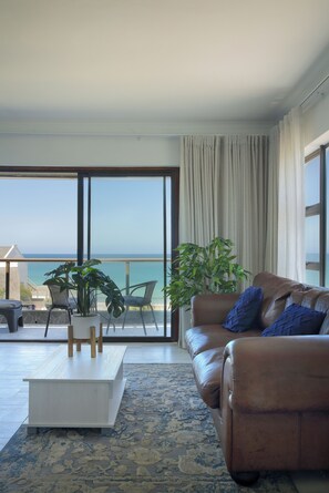 A living room with a large sliding glass door leading to a balcony with a view of the sea. There is a brown leather sofa with blue cushions, a white coffee table, and two chairs. A large potted plant is in the corner. - Mykonos Luxury Guest House (Saint Francis Bay)