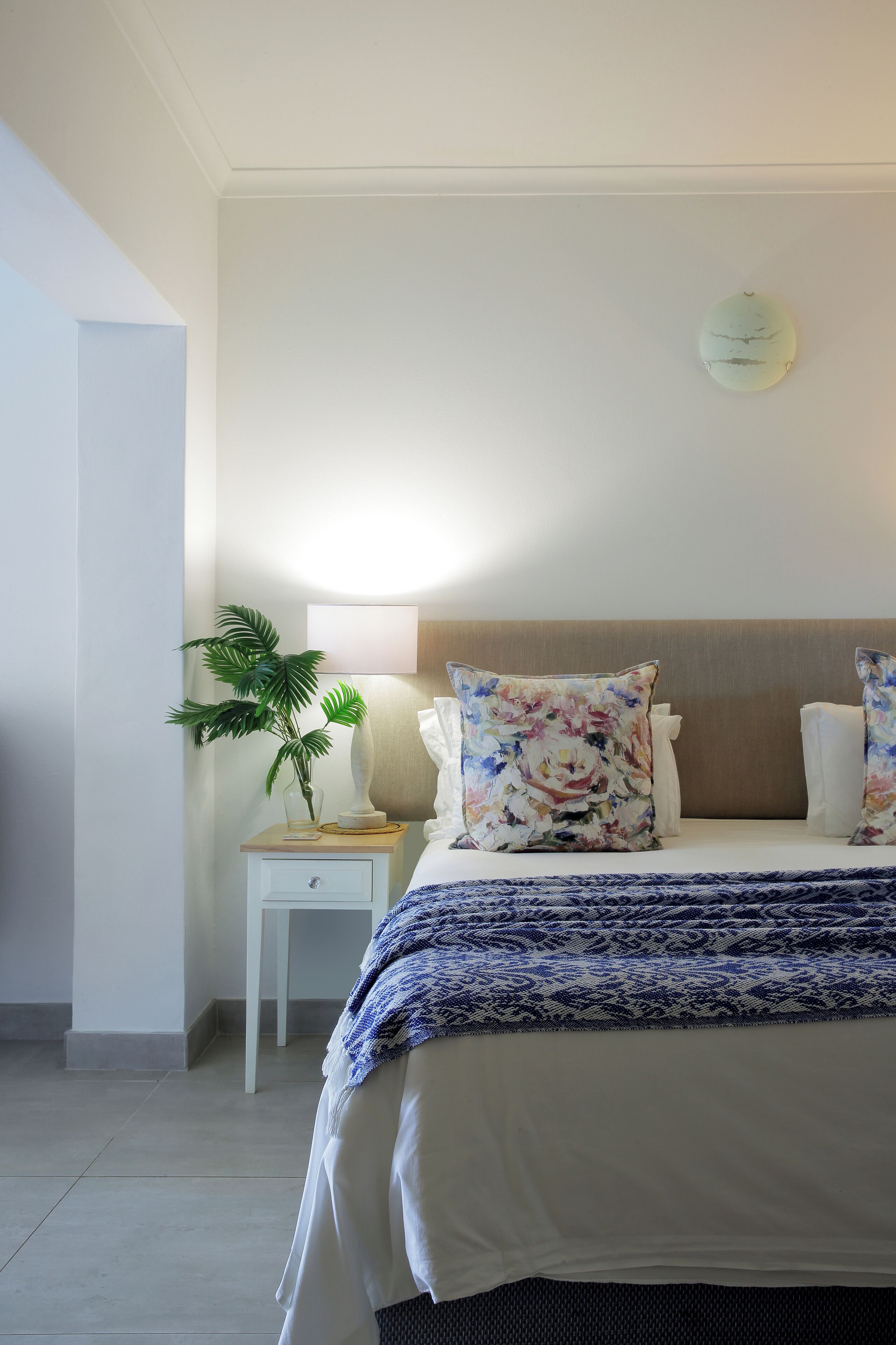 Un dormitorio ordenado con cama, mesita de noche con lámpara y planta en maceta.