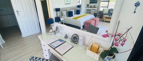 A hotel room with a large mirror, a desk with a chair, a bed with a yellow blanket, and a view of the outside through a window.