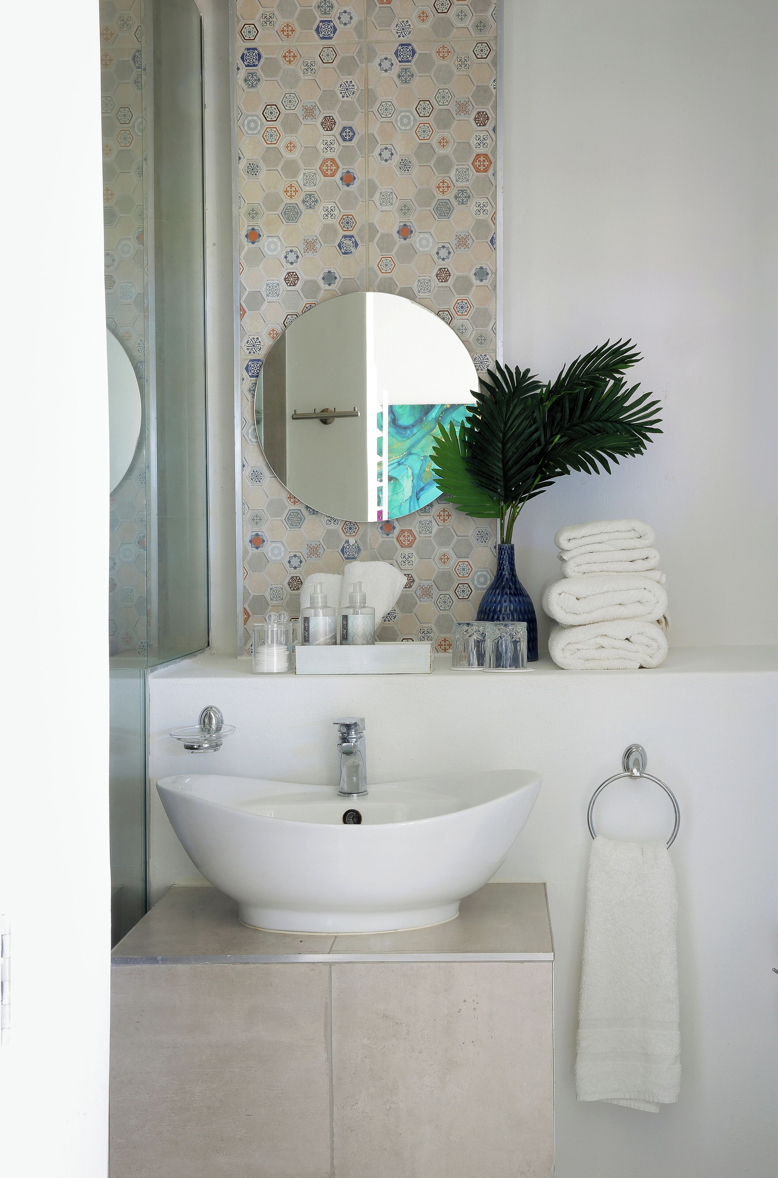 A bathroom with a white sink, a mirror, a blue vase with a plant, and a stack of white towels.