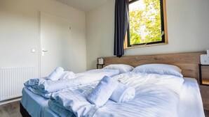 Un lit bien fait, avec du linge de lit blanc et des serviettes pliées, une tête de lit en bois, une table de chevet avec une lampe et une fenêtre donnant sur les arbres.