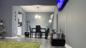 A modern living room with a grey carpet, a dining area with a black table and chairs, a mounted TV, and a large mirror. - Welcome To Your Comfy GetAway! (Dallas)