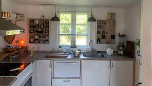 Una cocina con armarios blancos, una estufa, un fregadero y una ventana con persianas. Hay estantes con frascos y una licuadora en la encimera.