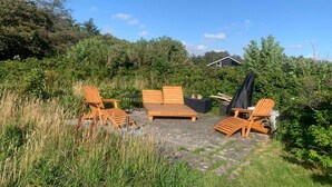 Un área de estar al aire libre con muebles de madera y suelo de piedra, rodeada de vegetación y una casa al fondo.