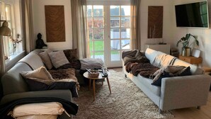 Una sala de estar con dos sofás, una mesa de centro y un televisor. Hay cortinas, una estatua de Buda y una planta.