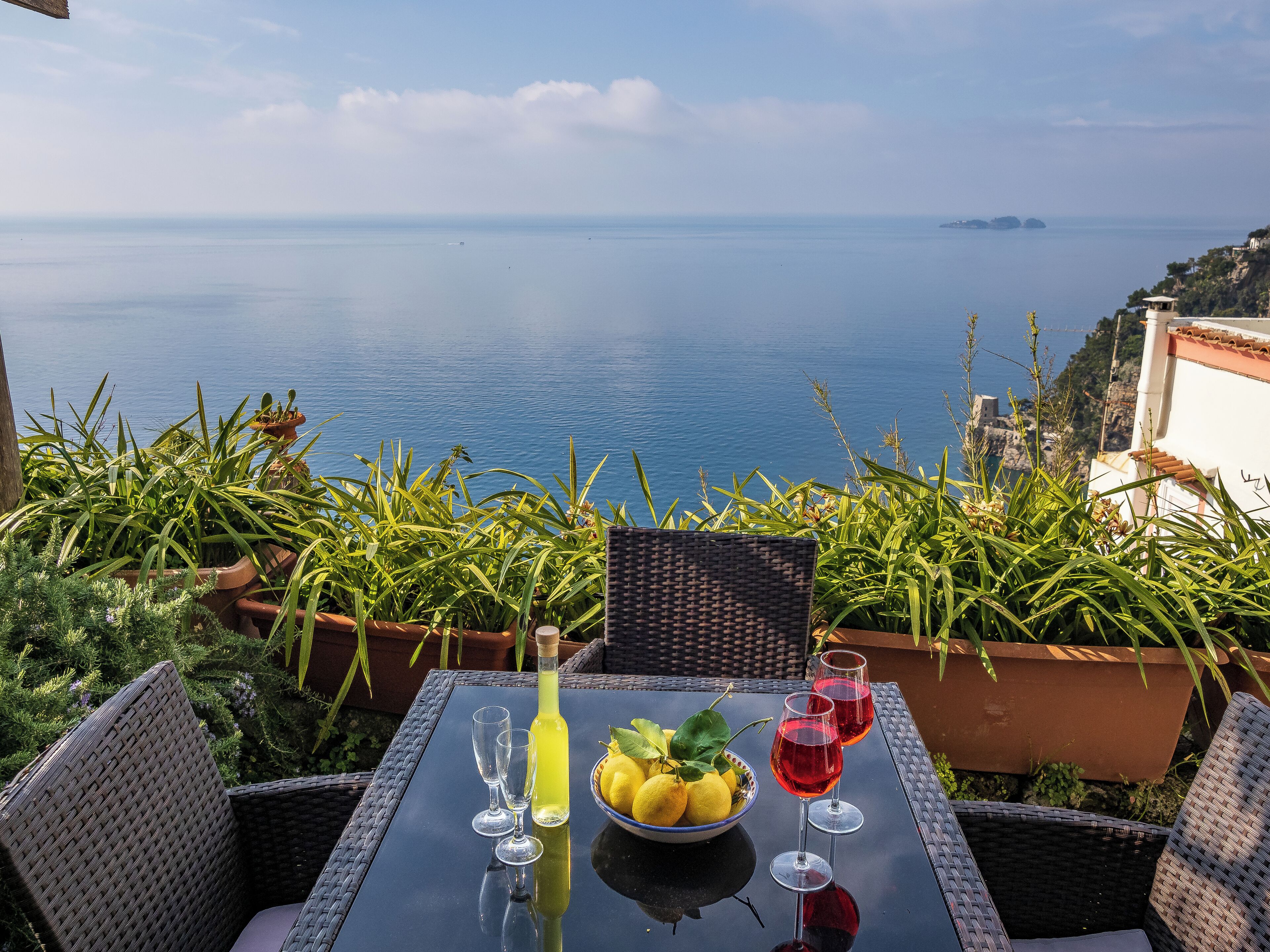 A table with a bottle, glasses, and a bowl of lemons and limes, set against a scenic ocean view.